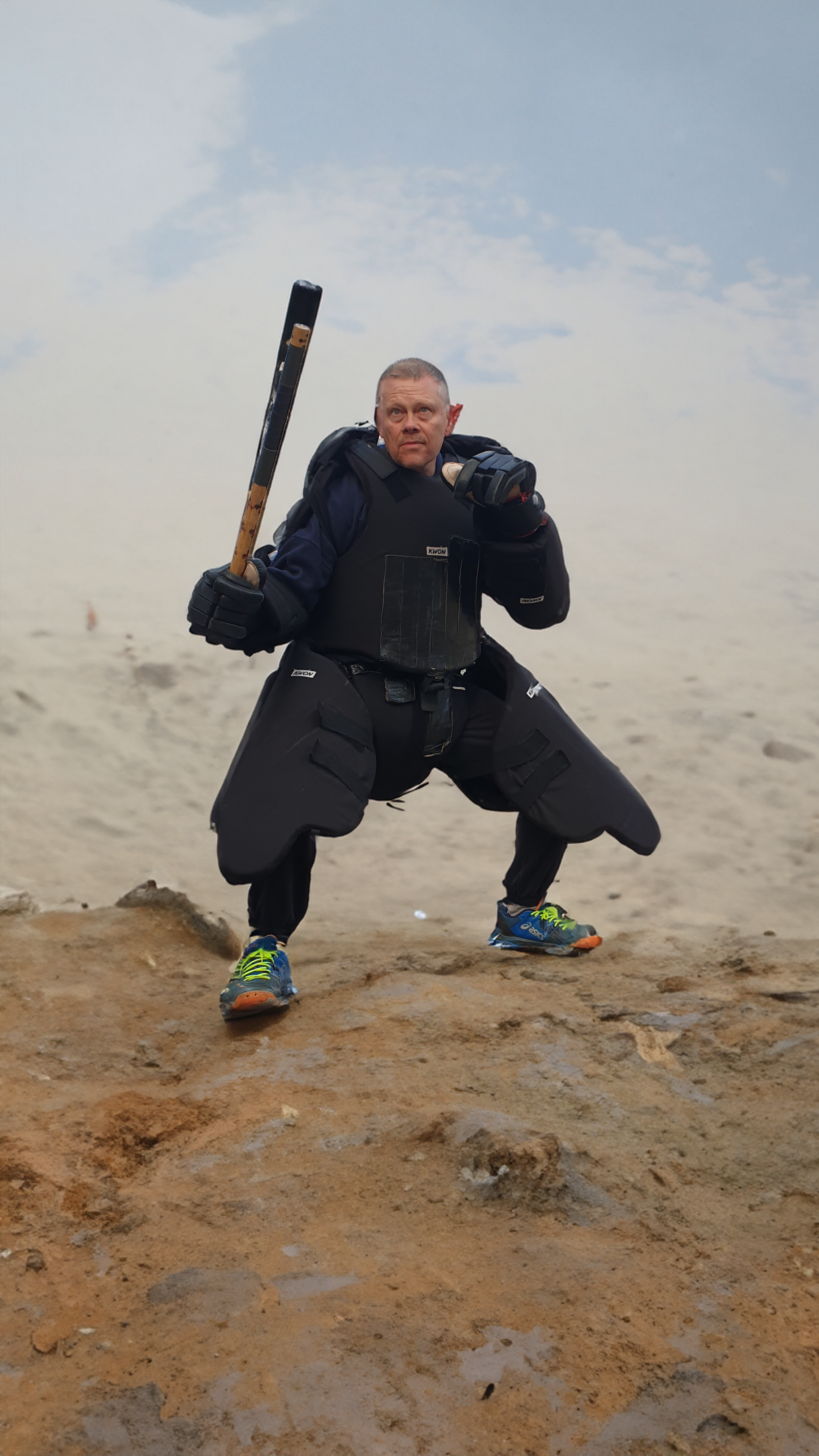 Oliver Solbach steht auf dem Gipfel eines hohen Berges. In seiner modernen Combat-Ausrüstung wirkt er wie ein unerschütterlicher Krieger. Der schwarze, taktische Kampfanzug ist mit Taschen und Polsterungen ausgestattet, und an seiner Hand hält er eine Waffe Mit einer stabilen Haltung und festem Blick auf die weite Landschaft wirkt er völlig fokussiert.