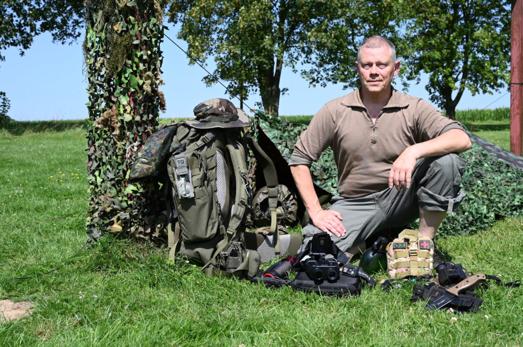 Oliver Solbach der auf dem Gras sitz und hinter ihm ein paar Bäume. Neben und vor ihm ist diverse Ausrüstung für das Outdoor Survival. man sieht Tarnausrüstung, Rucksack, Fernglas, Trinkflasche usw.