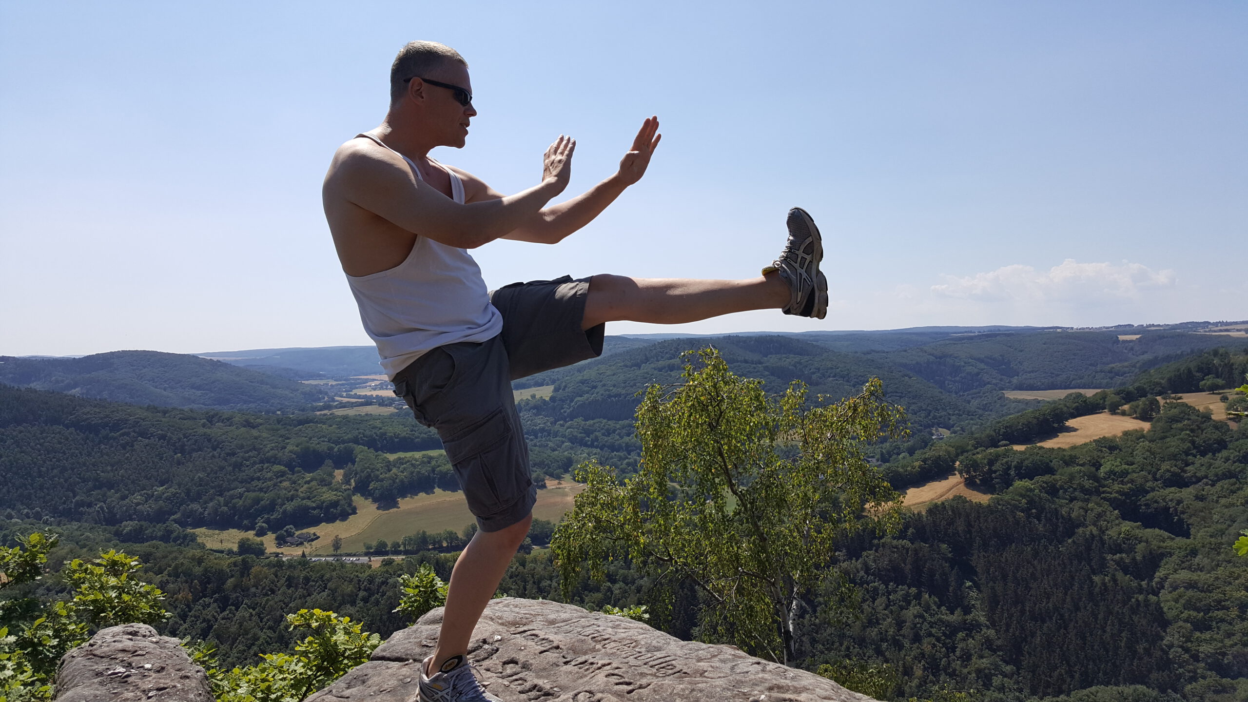 Oliver Solbach der auf der Bergspitze in der Combat-Outdoor-Training Logo Pose posiert. Im Hintergrund sieht man einen wunderschönen Wald.