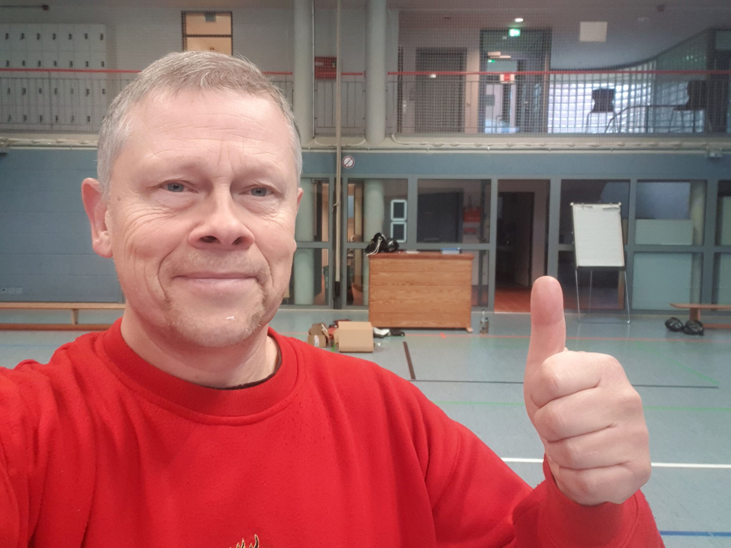 Oliver Solbach mit einem roten T-Shirt und den Daumen hoch. Im Hintergrund sieht man die Combat Trainingshalle, mit verschiedenen Trainingsgeräten.