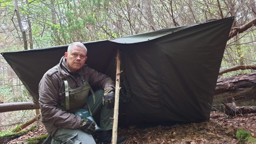 Oliver Solbach der für Outdoor Aktivitäten richtig gekleidet ist und sich im Wald befindet. Dabei hat er sich ein Unterschlupf gebaut mit einem langen Stock und einer Plane.