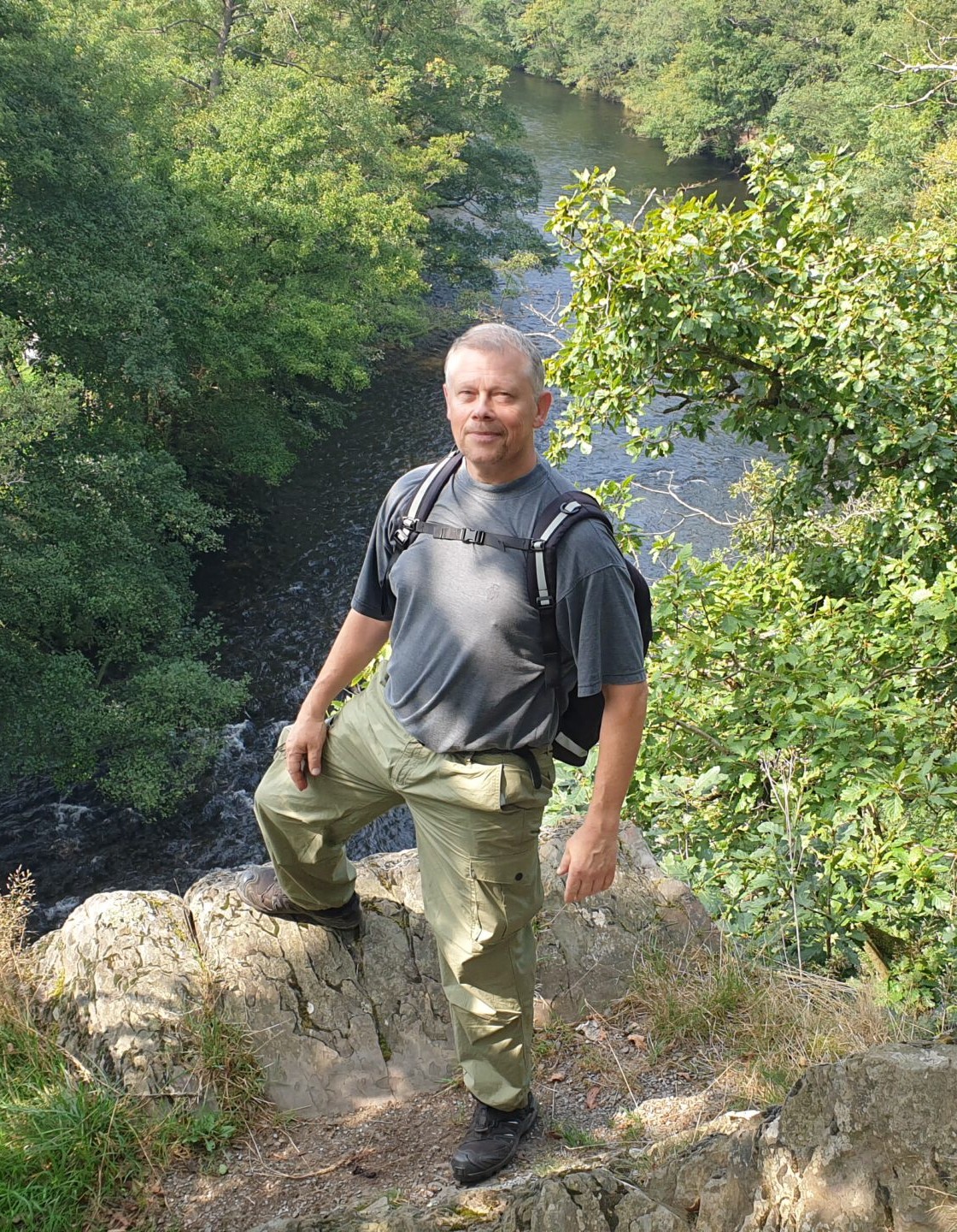 Oliver Solbach posiert im Outdoor auf einem Abhang. Im Hintergrund sieht man einen großen Wald der in der Mitte von einem Fluss geteilt wird.